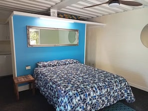 Une chambre à coucher avec un lit, un petit tabouret en bois, un miroir et un ventilateur de plafond.