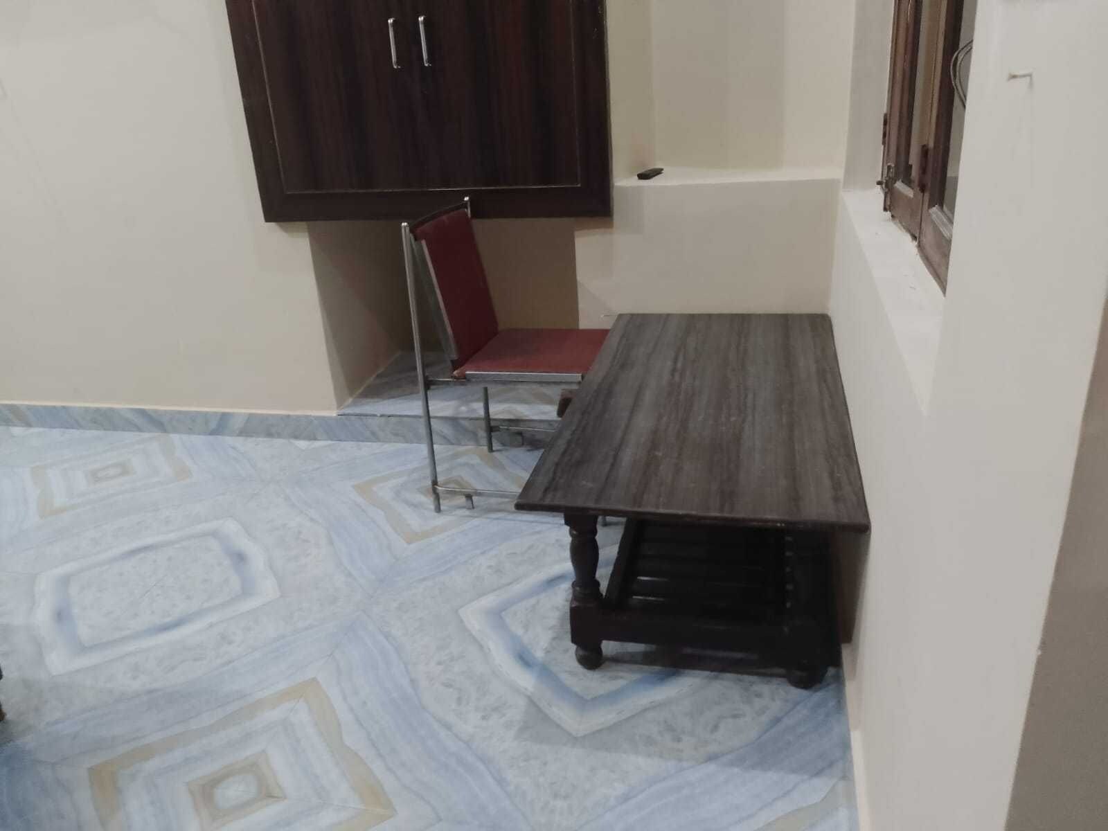 Una habitación pequeña con una mesa de madera oscura, una silla roja, una pared blanca y un suelo con diseño.