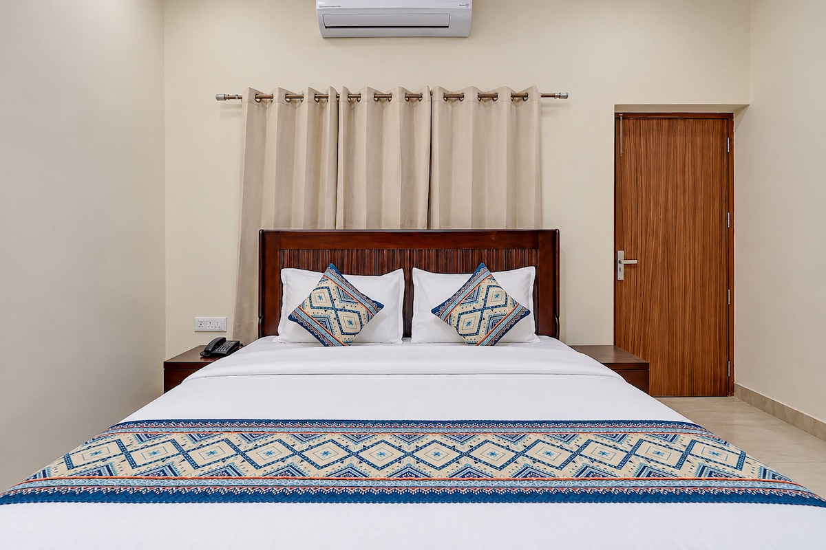 Une chambre d’hôtel avec un lit en bois, deux coussins à motifs, une couvre-lit blanche, une table de chevet en bois avec un téléphone, un rideau beige et une porte en bois.