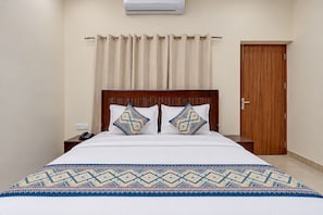Une chambre d’hôtel avec un lit en bois, deux coussins à motifs, une couvre-lit blanche, une table de chevet en bois avec un téléphone, un rideau beige et une porte en bois.