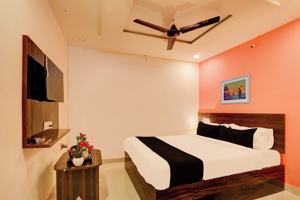 A hotel room with a bed, a ceiling fan, a wall-mounted TV, a small table with a vase of flowers, and a painting on the wall.