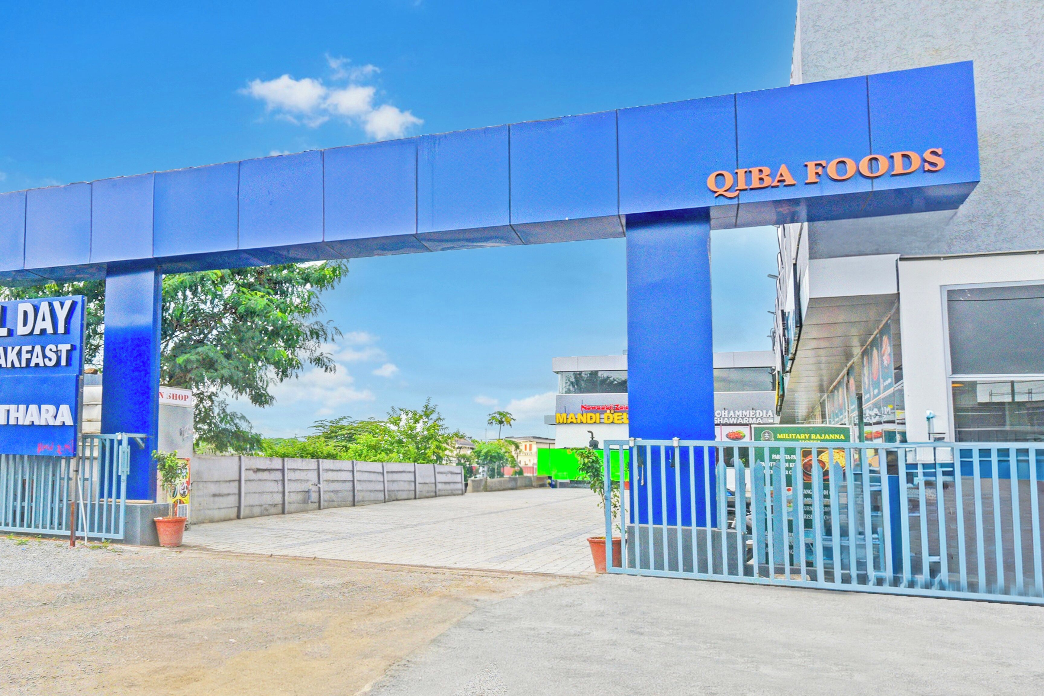 A gated entrance to a building with a blue canopy and signage for QIBA FOODS and a breakfast shop.