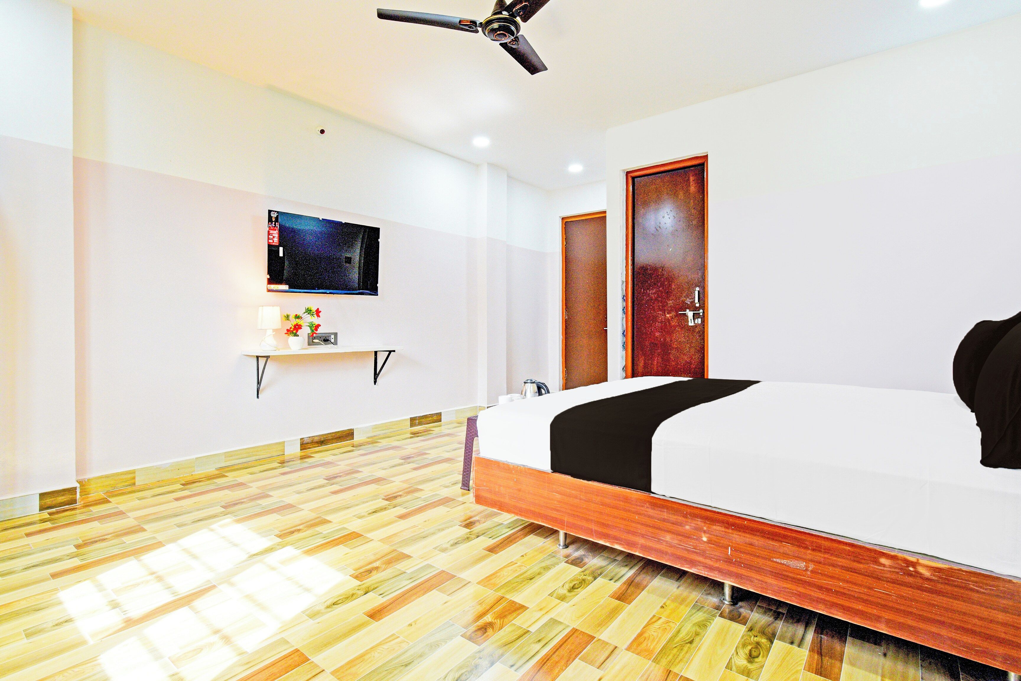 A hotel room with a bed, a flat-screen TV mounted on the wall, a wooden floor, and a ceiling fan.
