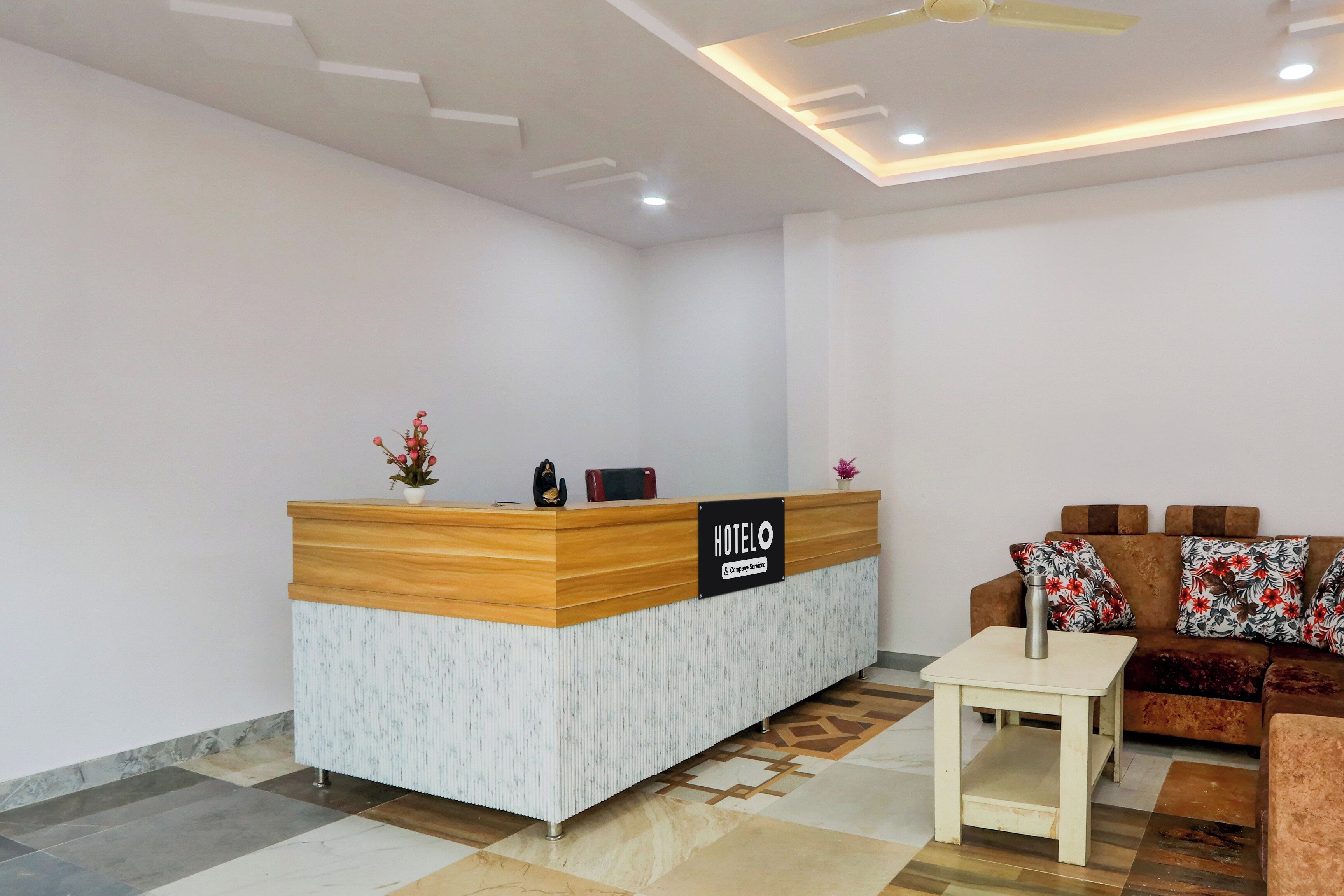 A hotel reception area with a wooden reception desk, a sofa with floral cushions, and a small table with a water bottle.