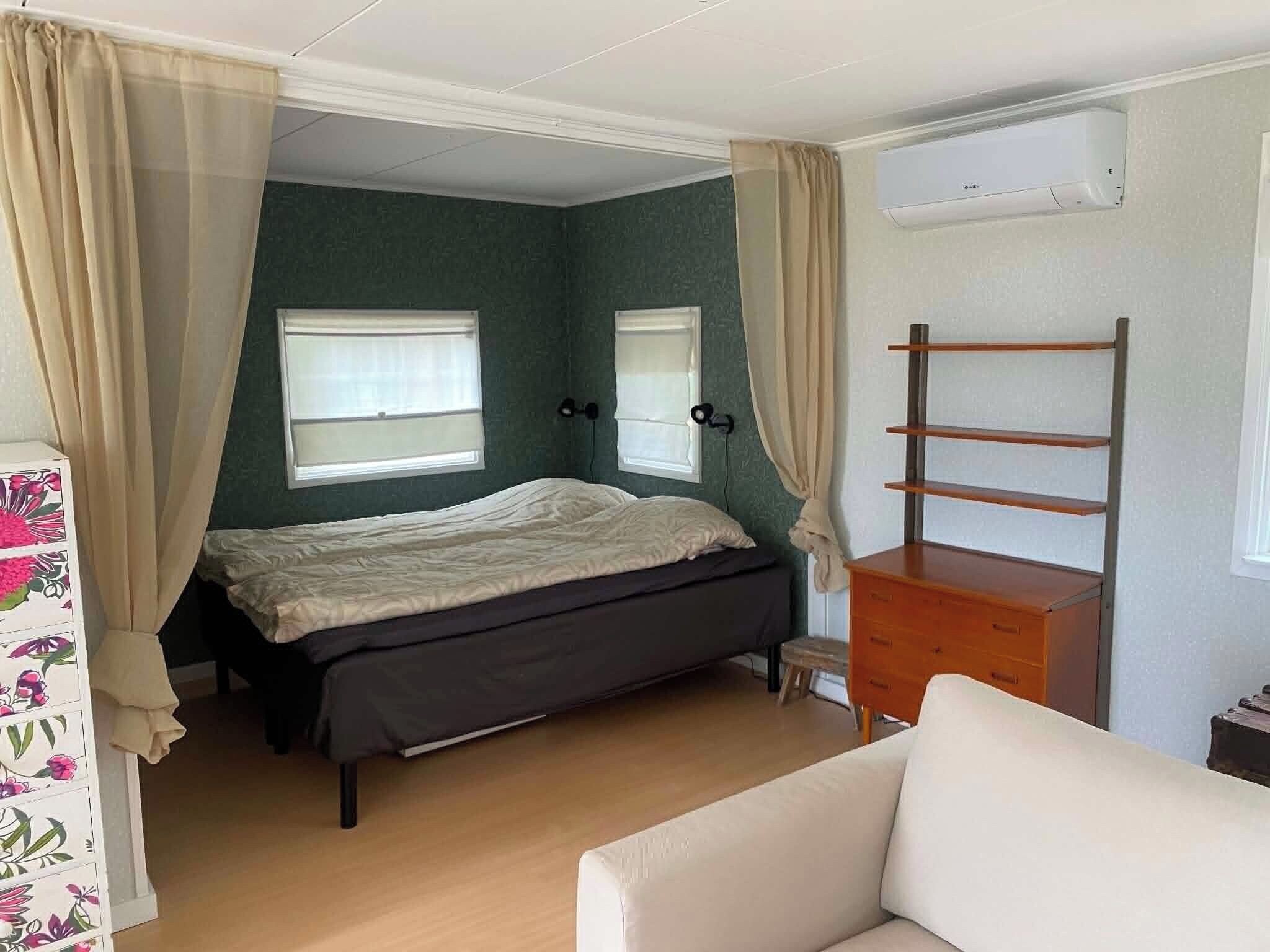 Un dormitorio con cama, cómoda, silla y dos ventanas con cortinas.