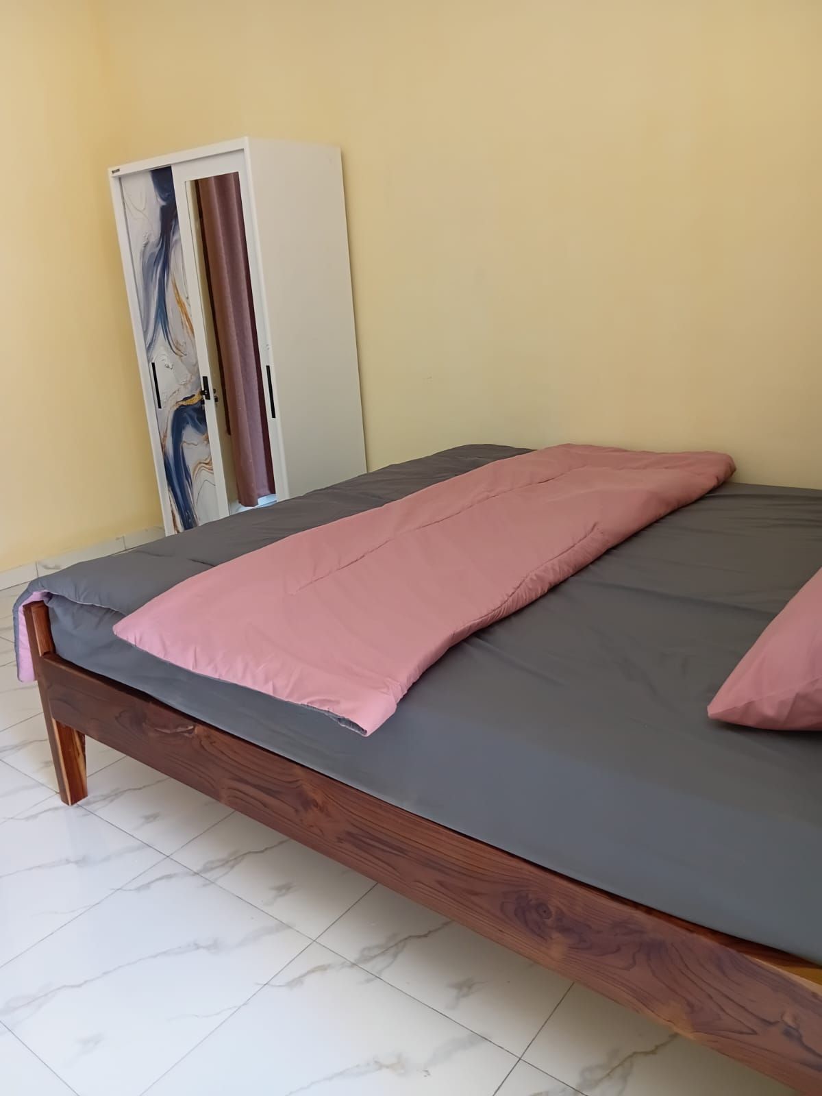 Una cama con colchón gris y ropa de cama rosa, un cabecero de madera y un armario con diseño de mármol.