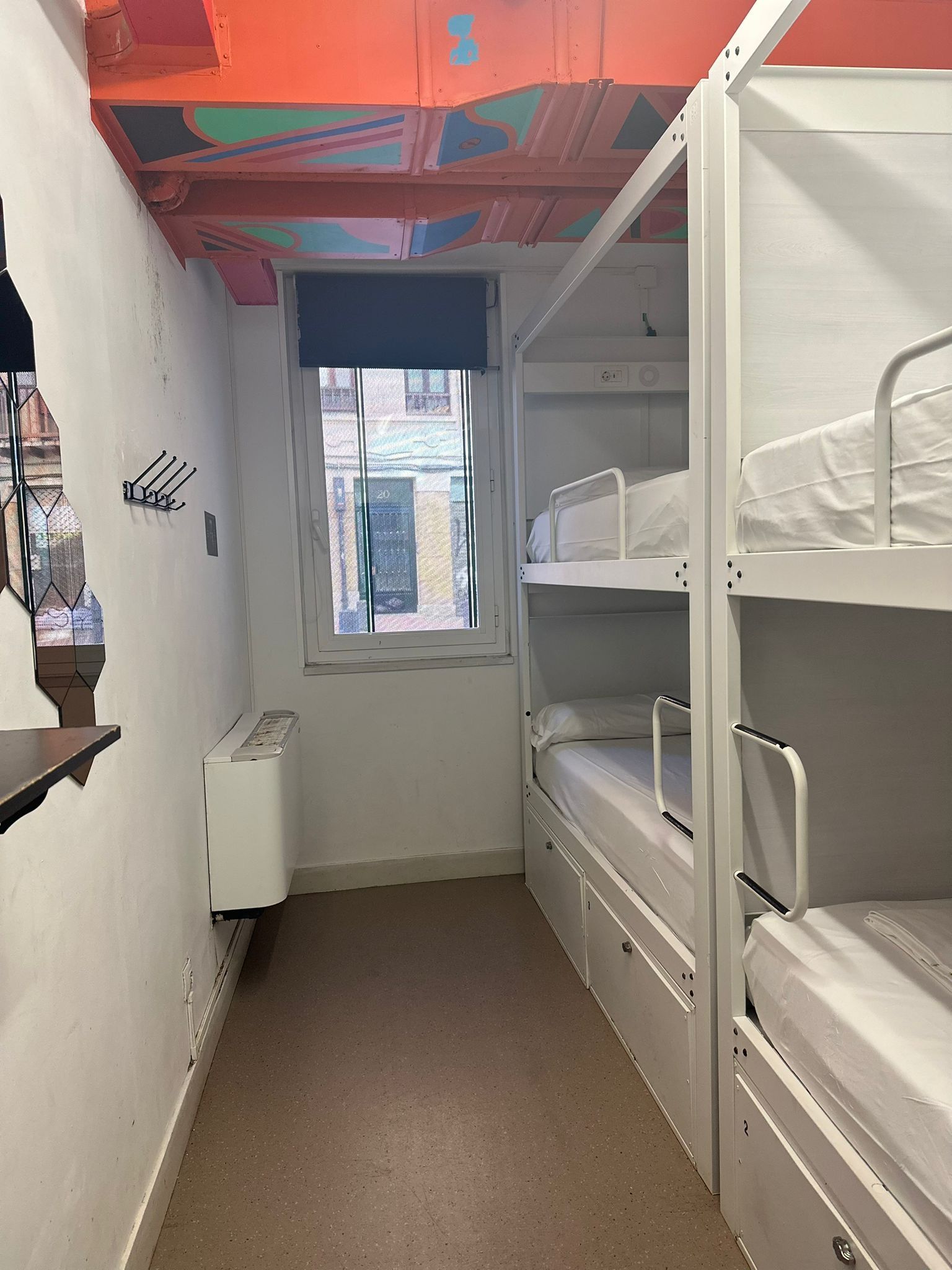 Habitación con literas, ventana, radiador y estante en la pared.