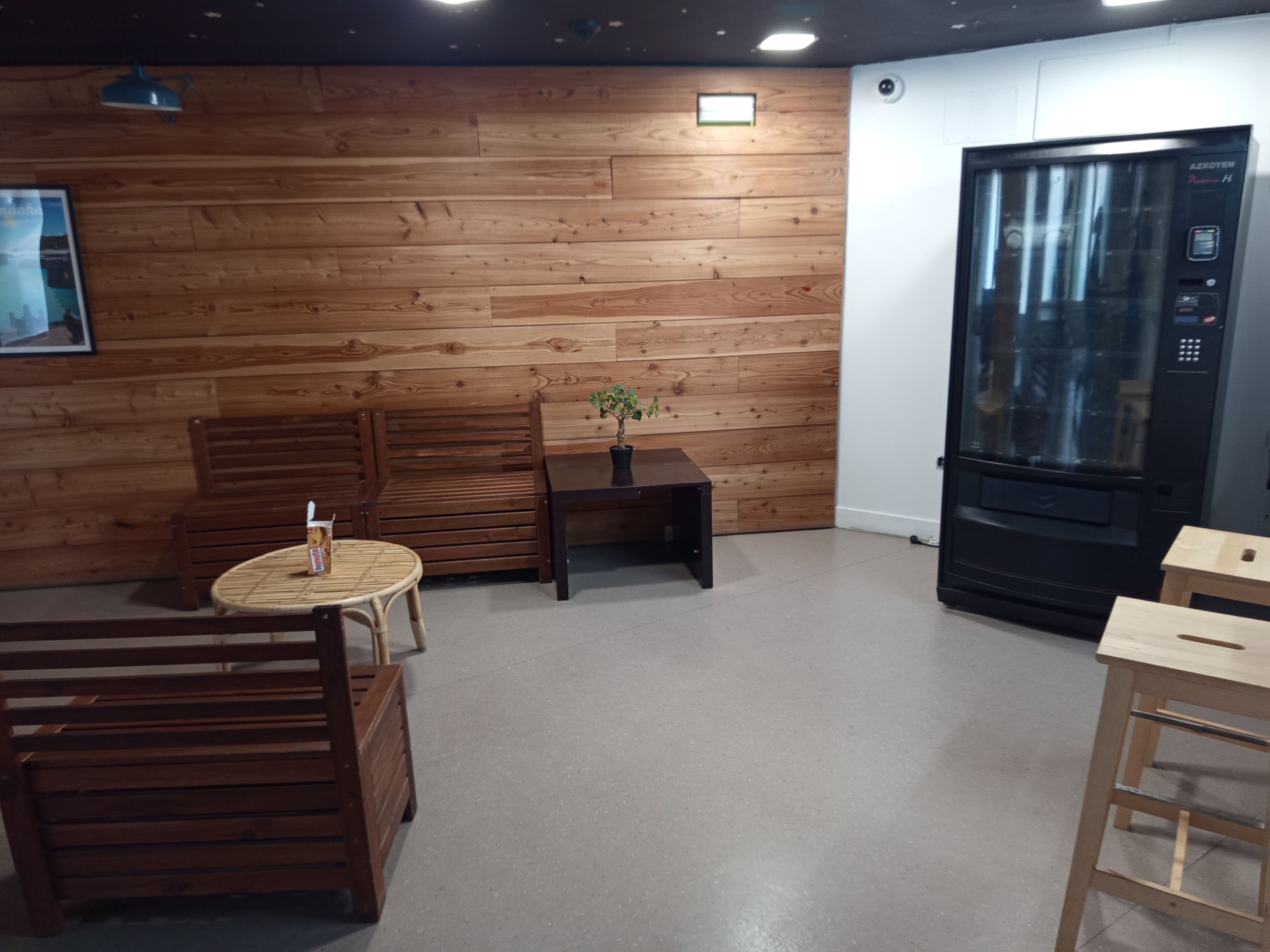 Een kamer met houten lambrisering, een houten bank, een tafeltje met een bloempot, een ronde tafel en een zwarte automaat.