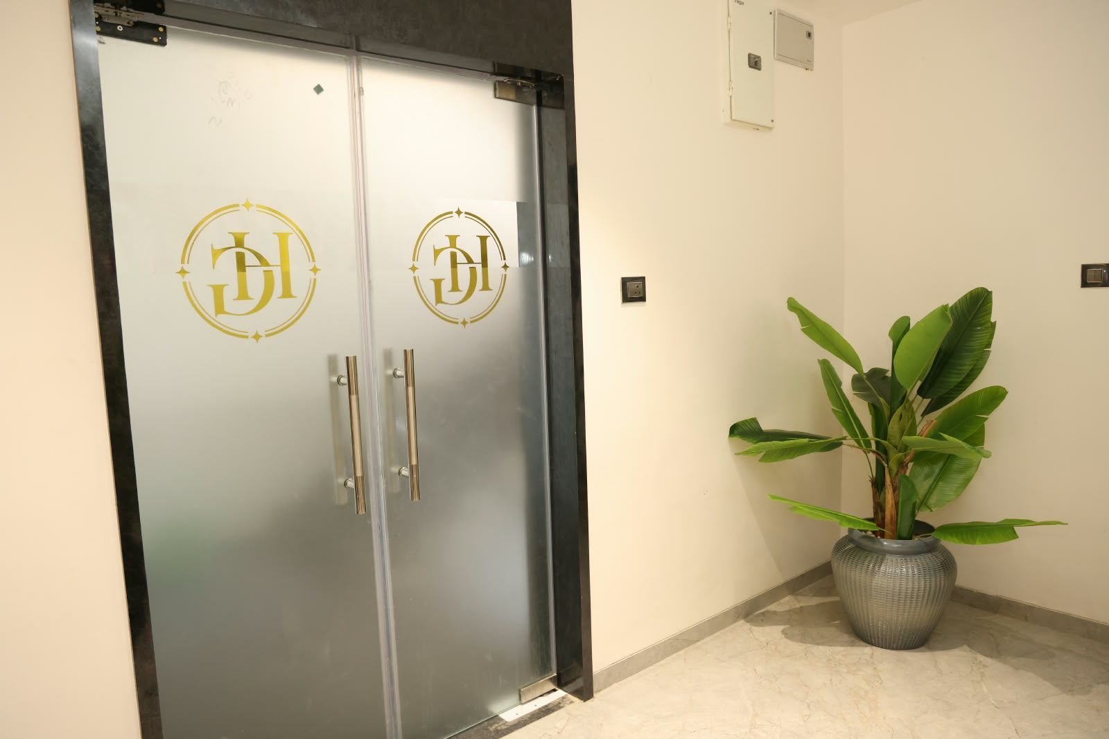 Unas puertas dobles con un logotipo, una planta en maceta y un interruptor de luz en la pared.