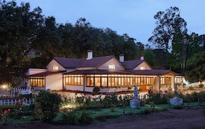 A large, two-story house with a wrap-around porch, surrounded by a well-maintained garden with various plants and flowers. - Miln Town by Zari (Coonoor)