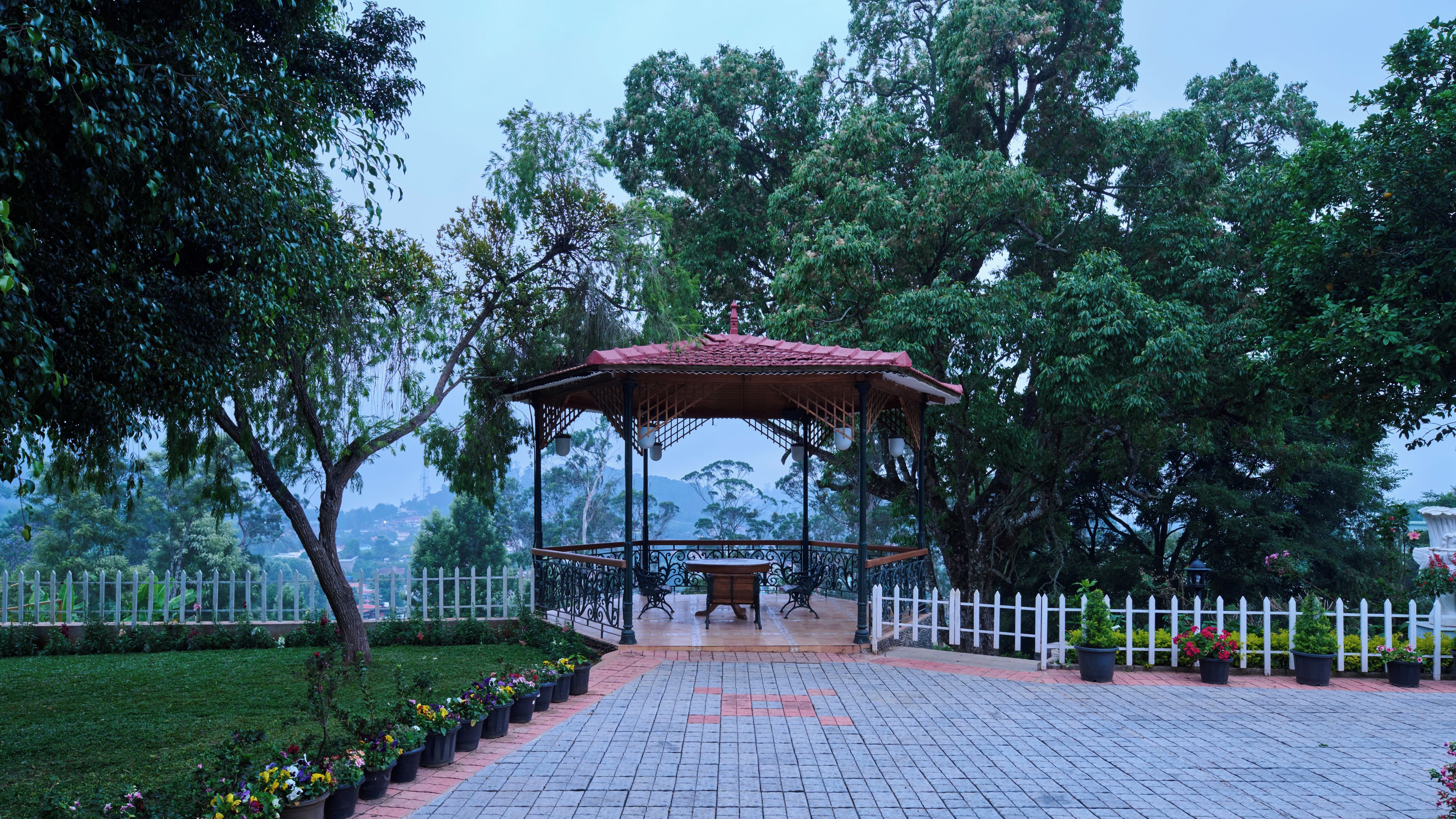 A gazebo with a tiled roof, surrounded by a white picket fence, set in a landscaped garden with potted plants and trees.