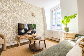 Un salon équipé d’un téléviseur à écran plat, d’une table basse ronde en bois, d’un canapé, d’une grande fenêtre donnant sur des immeubles et d’une plante en pot.