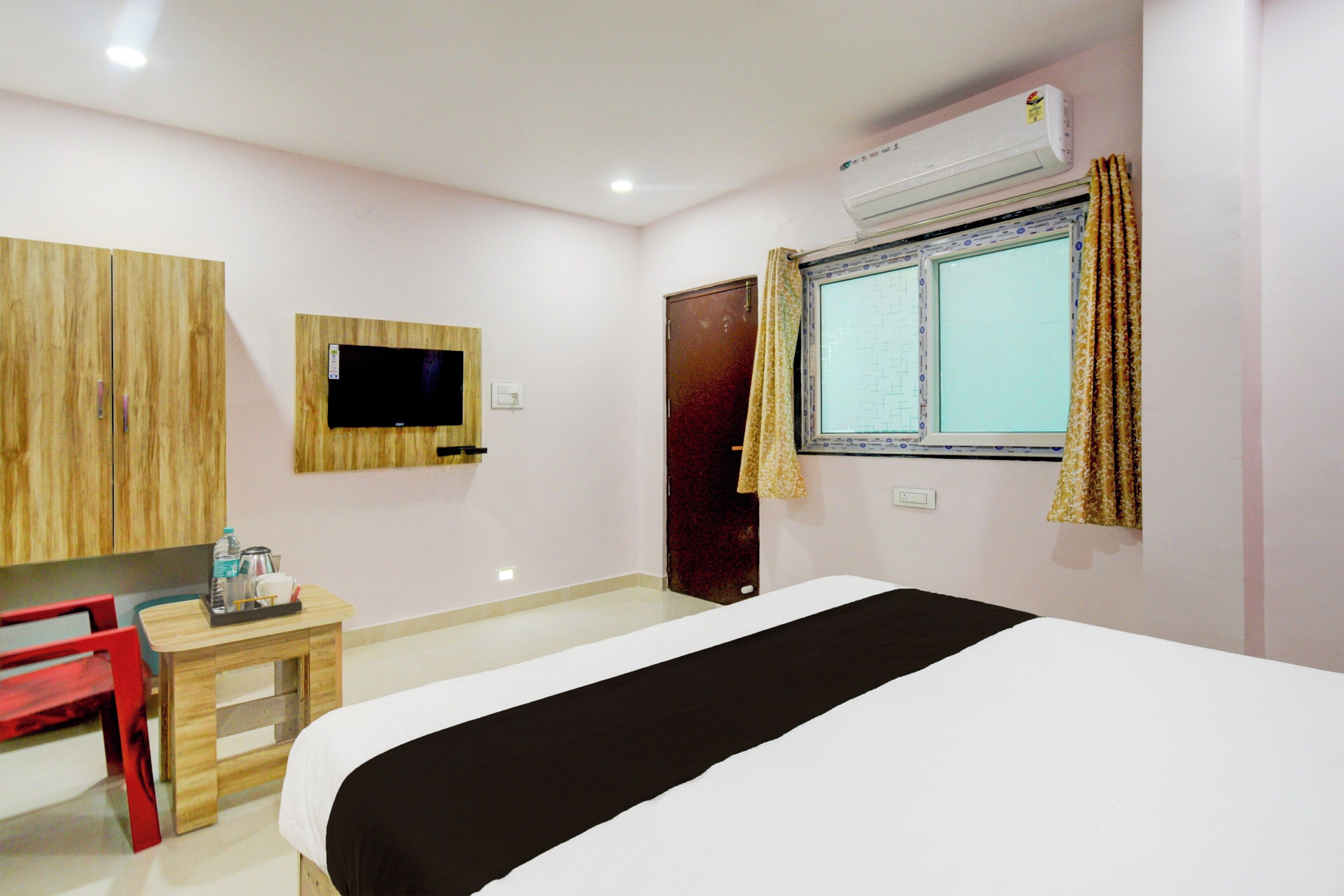 A hotel room with a bed, a television mounted on the wall, a small table with a chair, and a window with curtains.