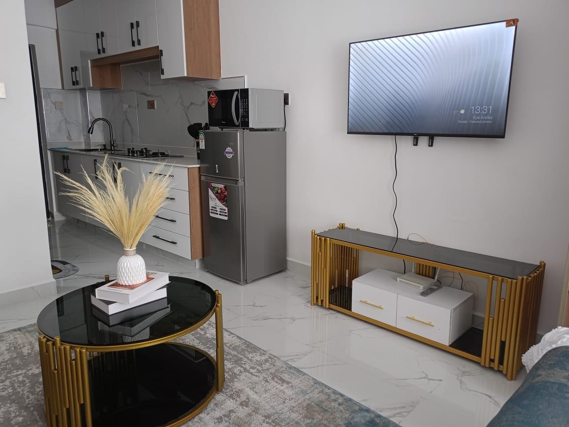 Een moderne woonkamer met een flatscreen televisie aan de muur, een glazen salontafel met een decoratieve vaas, een witte bank en een keuken met een koelkast en magnetron.