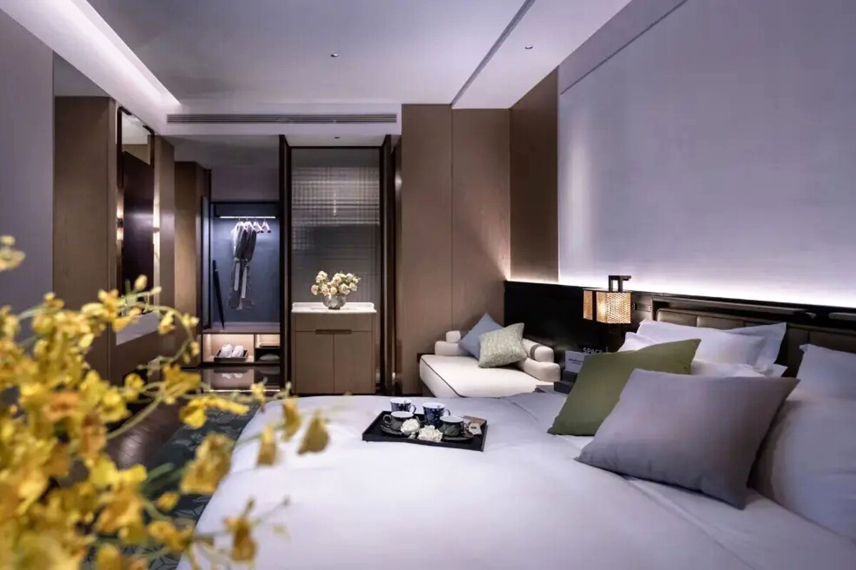 Quarto de hotel moderno com uma cama grande, área de estar e vista para um edifício.