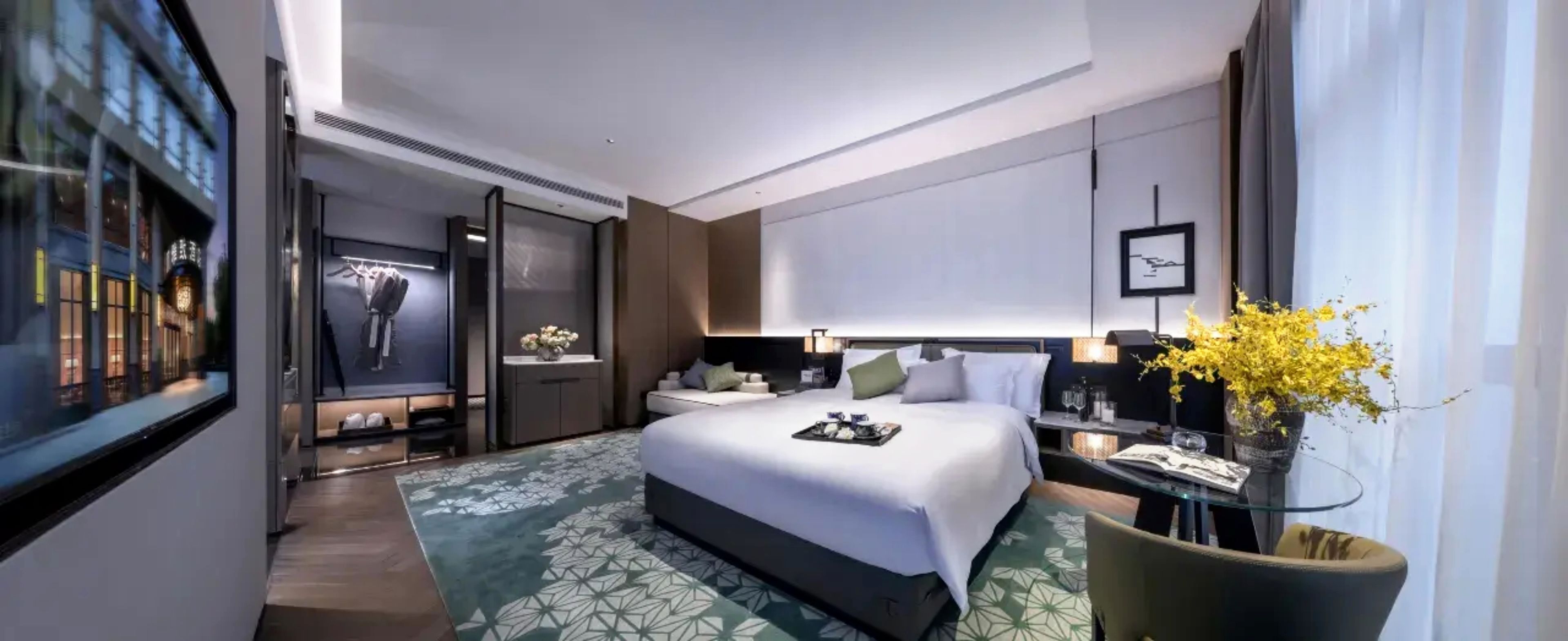 A modern hotel room with a large bed, bedside tables, a desk, and a view of the city.