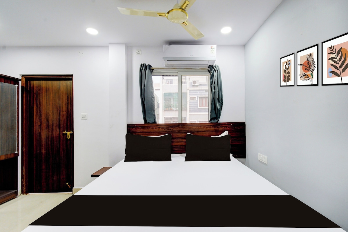 A hotel room with a bed, a ceiling fan, an air conditioning unit, a window with curtains, and framed artwork on the wall.