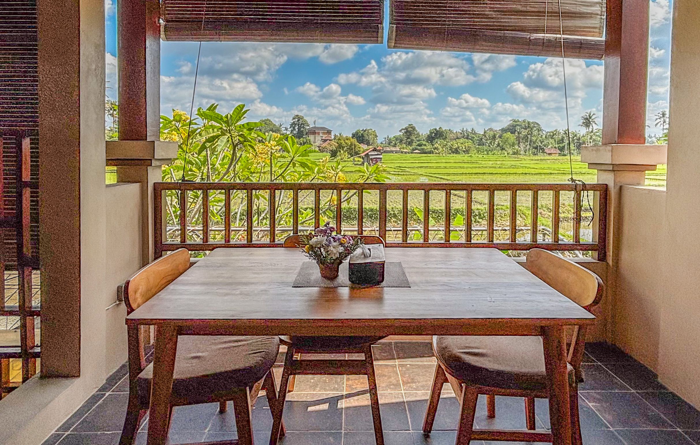A wooden table with four chairs and a vase of flowers on a balcony with a view of a green field and a house.