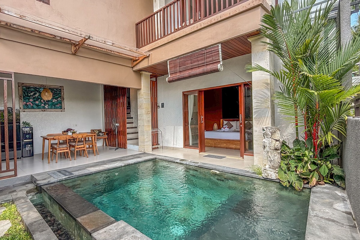 A two-story building with a swimming pool, wooden furniture, and tropical plants.
