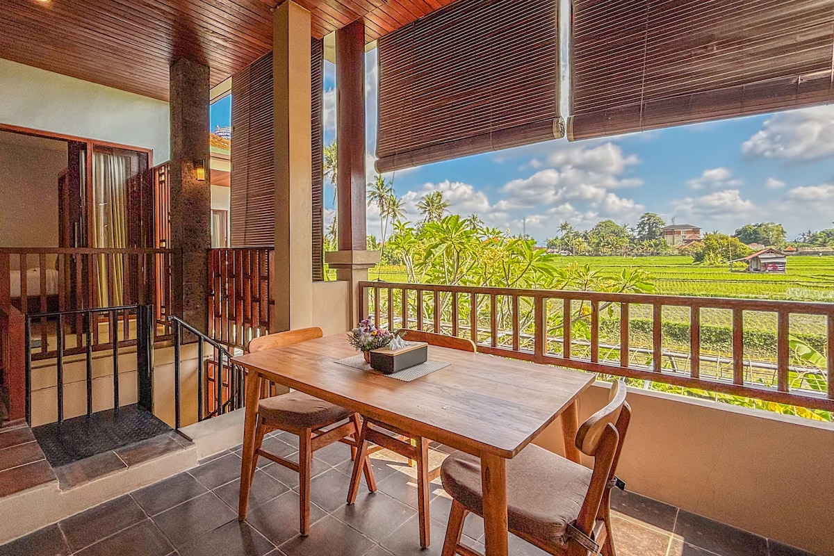 A wooden dining table with chairs, a view of a green field, and a balcony railing.
