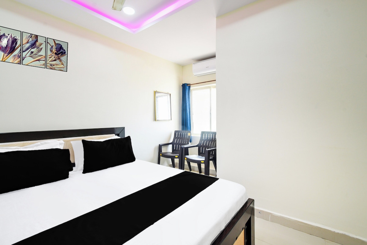 Habitación de hotel con cama, escritorio, dos sillas, ventana con cortinas y lámpara en el techo.