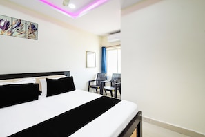 Habitación de hotel con cama, escritorio, dos sillas, ventana con cortinas y lámpara en el techo.