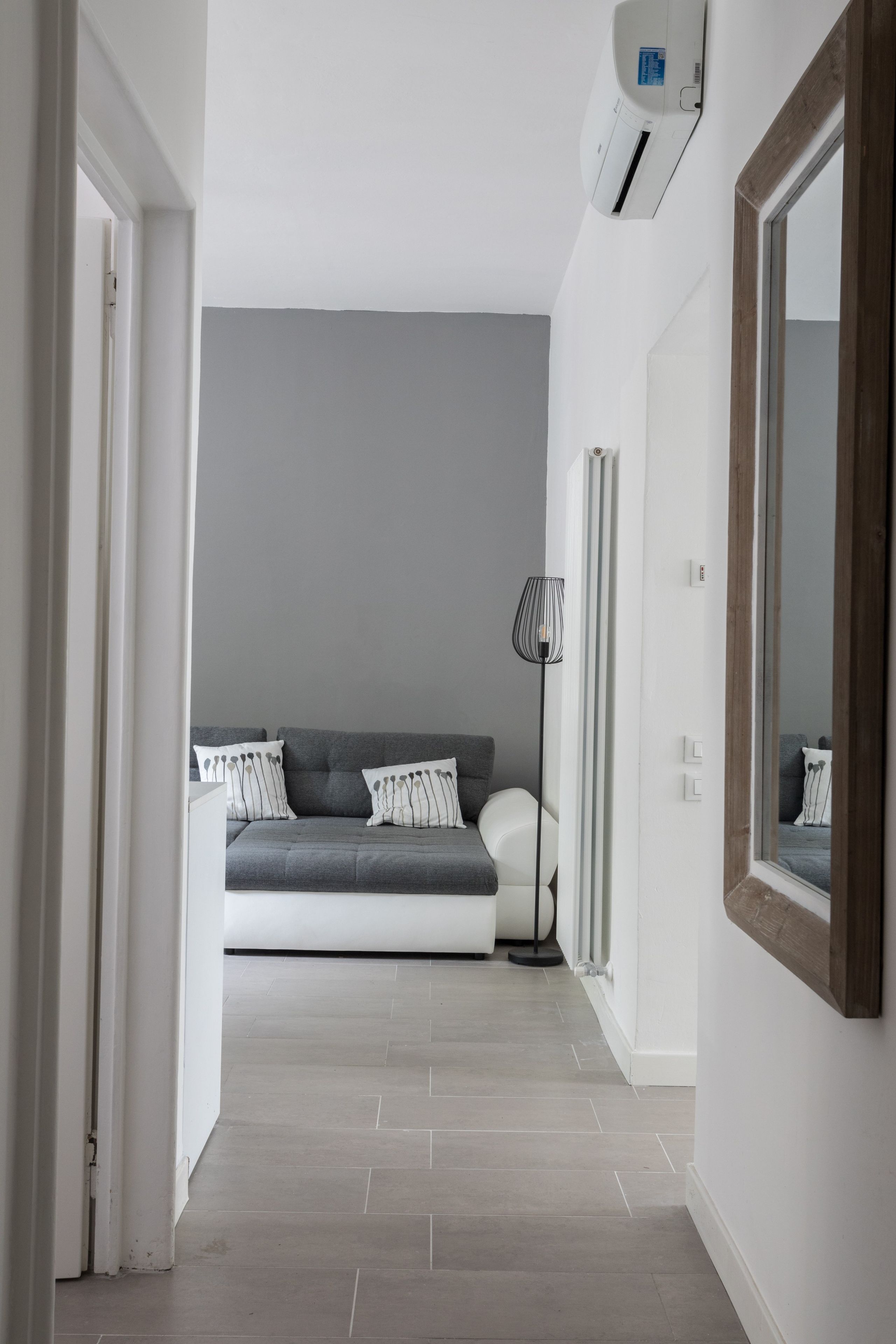 Ein Flur, der zu einem Schlafzimmer mit grauem Sofa, Stehlampe und großem Spiegel führt.