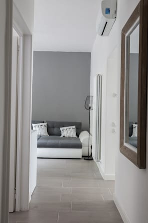 Ein Flur, der zu einem Schlafzimmer mit grauem Sofa, Stehlampe und großem Spiegel führt.