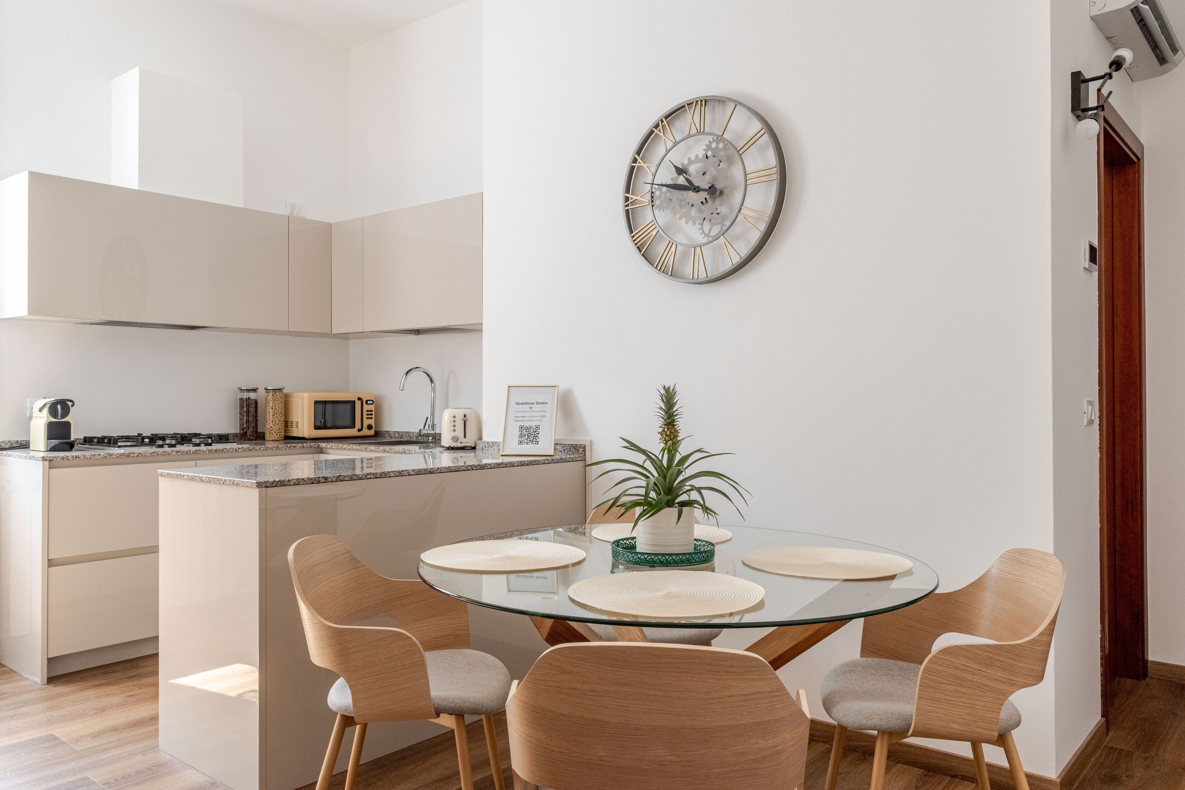 Eine moderne Küche mit Mikrowelle, Toaster und Wasserkocher. Es gibt eine Essecke mit einem Glastisch und vier Stühlen. Eine dekorative Uhr befindet sich an der Wand.