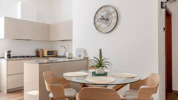 A modern kitchen with a microwave, toaster, and kettle. There is a dining area with a glass table and four chairs. A decorative clock is on the wall.