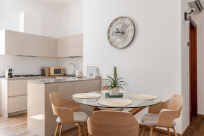 Eine moderne Küche mit Mikrowelle, Toaster und Wasserkocher. Es gibt eine Essecke mit einem Glastisch und vier Stühlen. Eine dekorative Uhr befindet sich an der Wand.