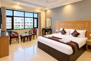 A hotel room with a large bed, a desk, two chairs, a window with a view of buildings, and a ceiling fan. - Eva Hotel (Zirakpur)
