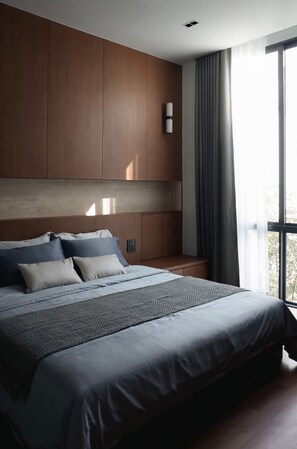 Une chambre à coucher moderne avec un grand lit, une tête de lit en bois et une vue sur l’extérieur par une fenêtre.