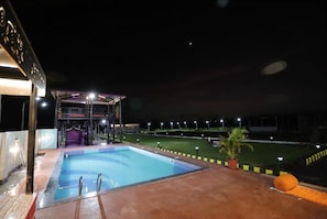 Una piscina con zona cubierta, un campo deportivo vallado y un edificio con balcón.