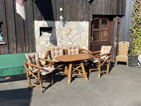 Un espace extérieur aménagé avec des meubles en bois, comprenant une table ronde et six chaises, un fauteuil en osier et un mur en pierre en arrière-plan.