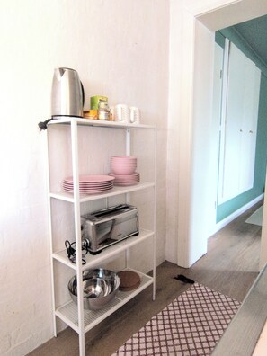 Cozinha com prateleira metálica branca contendo pratos cor-de-rosa, chaleira prateada e torradeira.