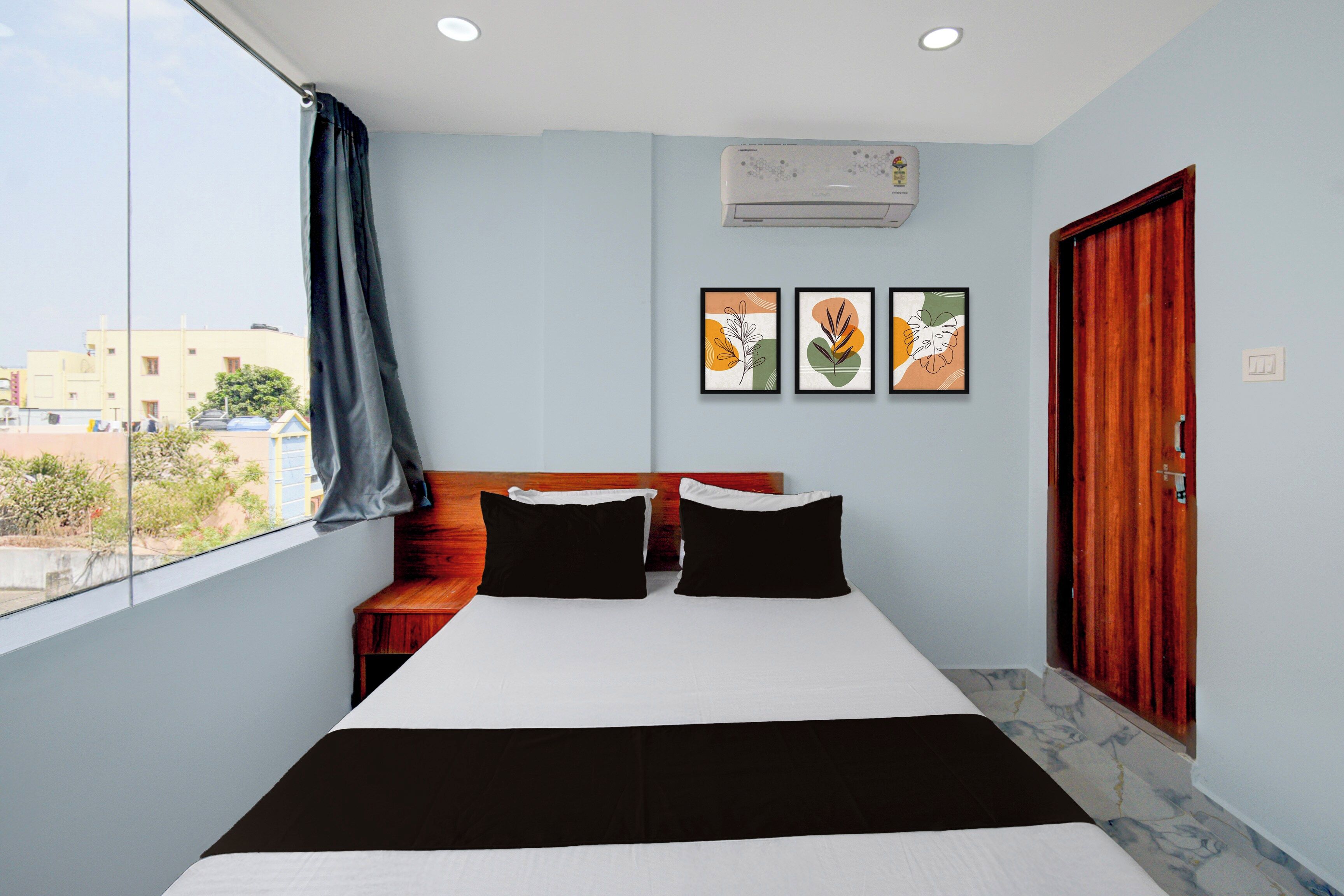 A bedroom with a bed, two pillows, a wooden headboard, a window with a view of buildings, and three framed artworks on the wall.