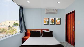 A bedroom with a bed, two pillows, a wooden headboard, a window with a view of buildings, and three framed artworks on the wall.