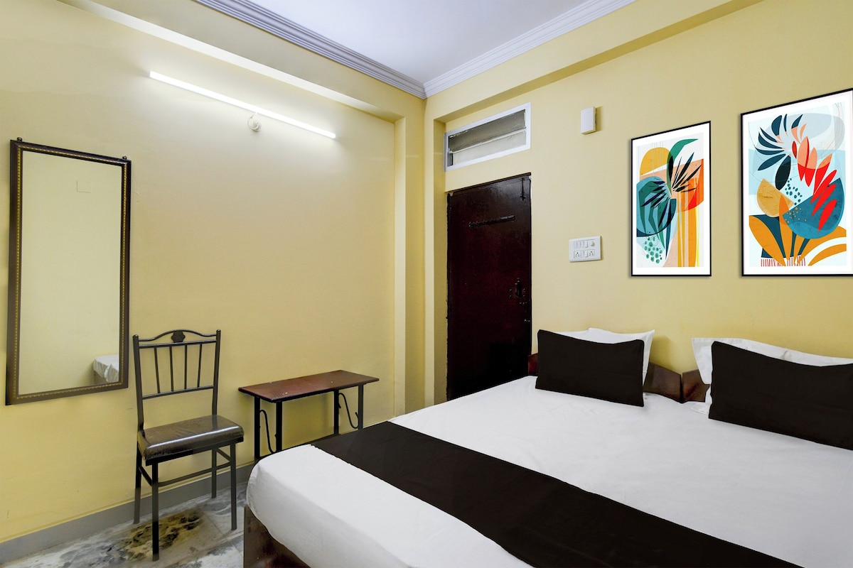 A hotel room with a bed, a chair, a small table, a mirror, and two abstract paintings on the wall.
