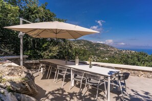 An outdoor dining area with a table and chairs, a large umbrella, and a scenic view of a coastal landscape.