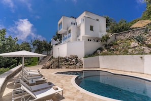 A modern two-story house with a swimming pool, outdoor seating, and a stone retaining wall.