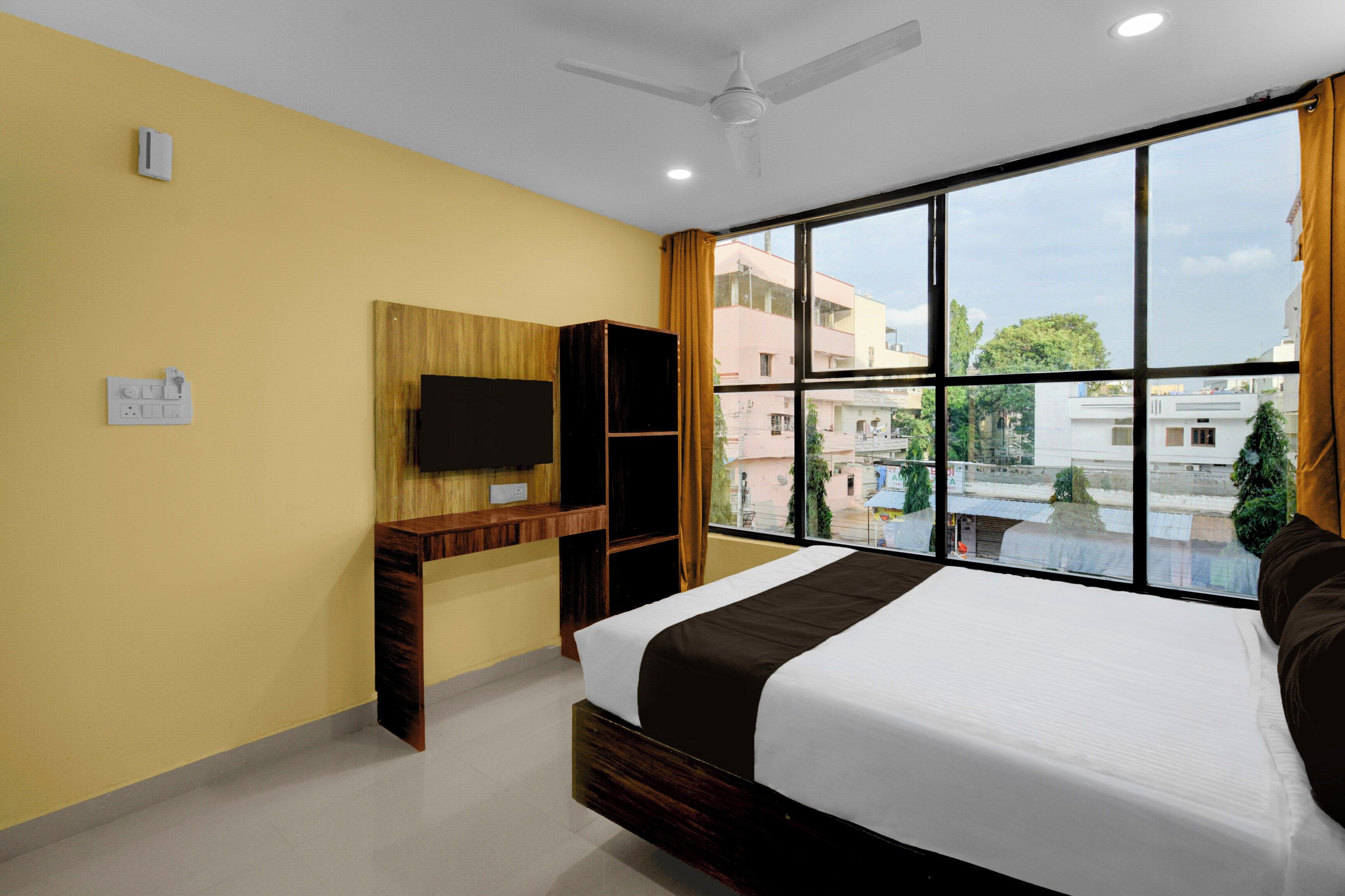 A hotel room with a bed, a TV mounted on the wall, a desk, and a window with a view of buildings.