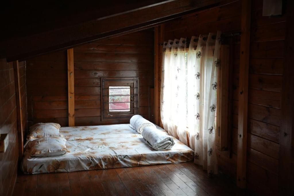 Une petite pièce aux murs en bois, faiblement éclairée, avec un lit simple recouvert d’une courtepointe à motifs floraux et une fenêtre ornée d’un rideau.