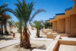 Un resort al desert amb palmeres, una platja sorrenca i un edifici amb teulada plana i parets beix.