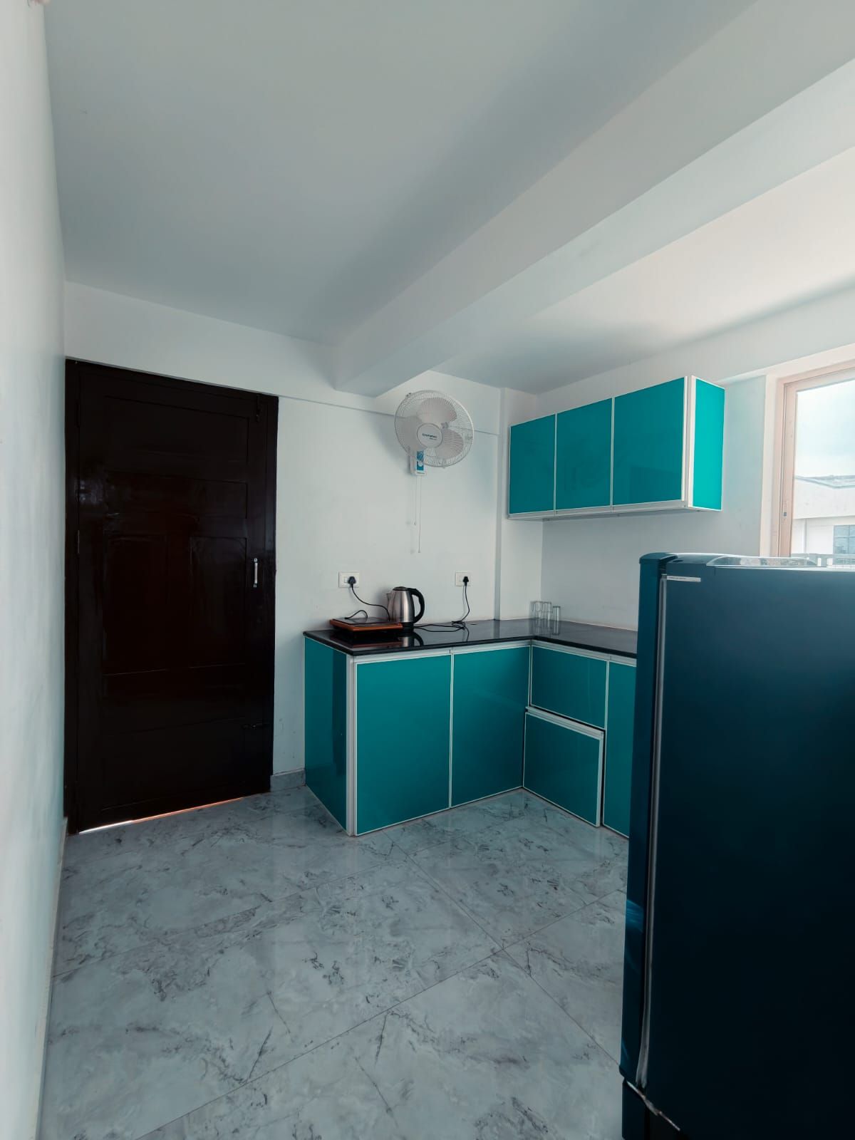 Una cocina compacta con armarios color turquesa, un refrigerador negro y un ventilador de techo blanco.