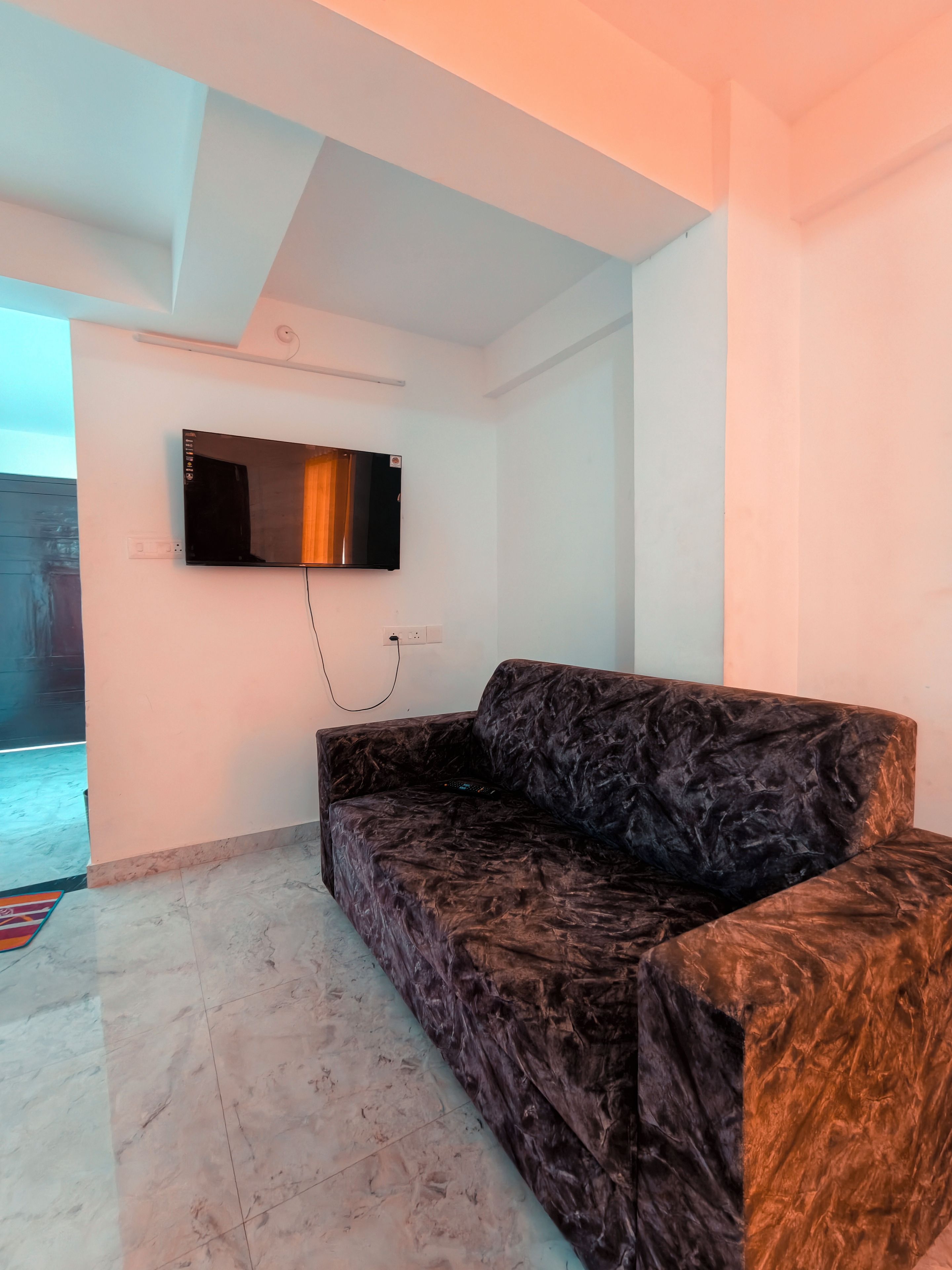 A room with a dark patterned sofa, a flat-screen TV mounted on the wall, and a tiled floor.