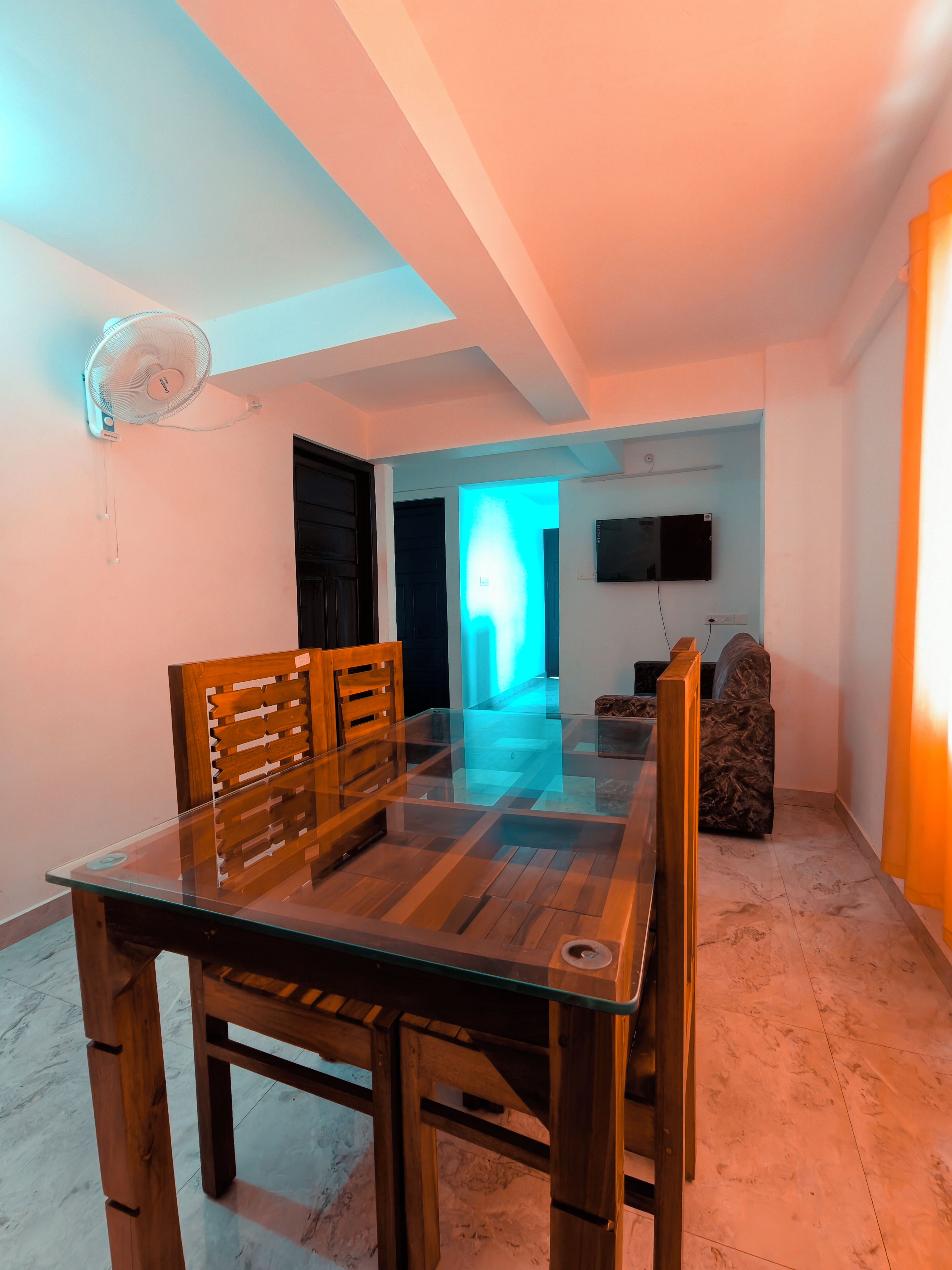 A dining area with a glass table and wooden chairs, a ceiling fan, and a television on the wall.