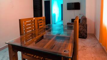 A dining area with a glass table and wooden chairs, a ceiling fan, and a television on the wall.