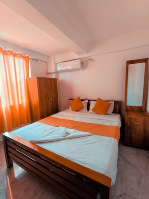 A bedroom with a bed, wooden headboard, orange pillows, a bedside table, a wardrobe, and an air conditioning unit. - inn city apartments (Kazhakkoottam)