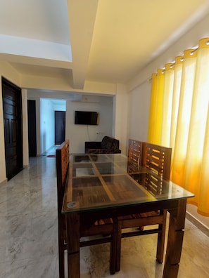 A dining area with a glass-top table, wooden chairs, a flat-screen TV mounted on the wall, and a yellow curtain. - inn city apartments (Kazhakkoottam)